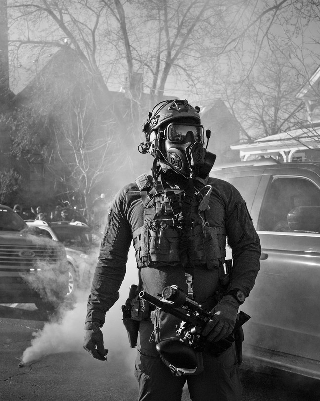Black-and-white photo of a federal agent seen on a Minneapolis neighborhood street wearing a gas mask while tear gas goes off in the background.