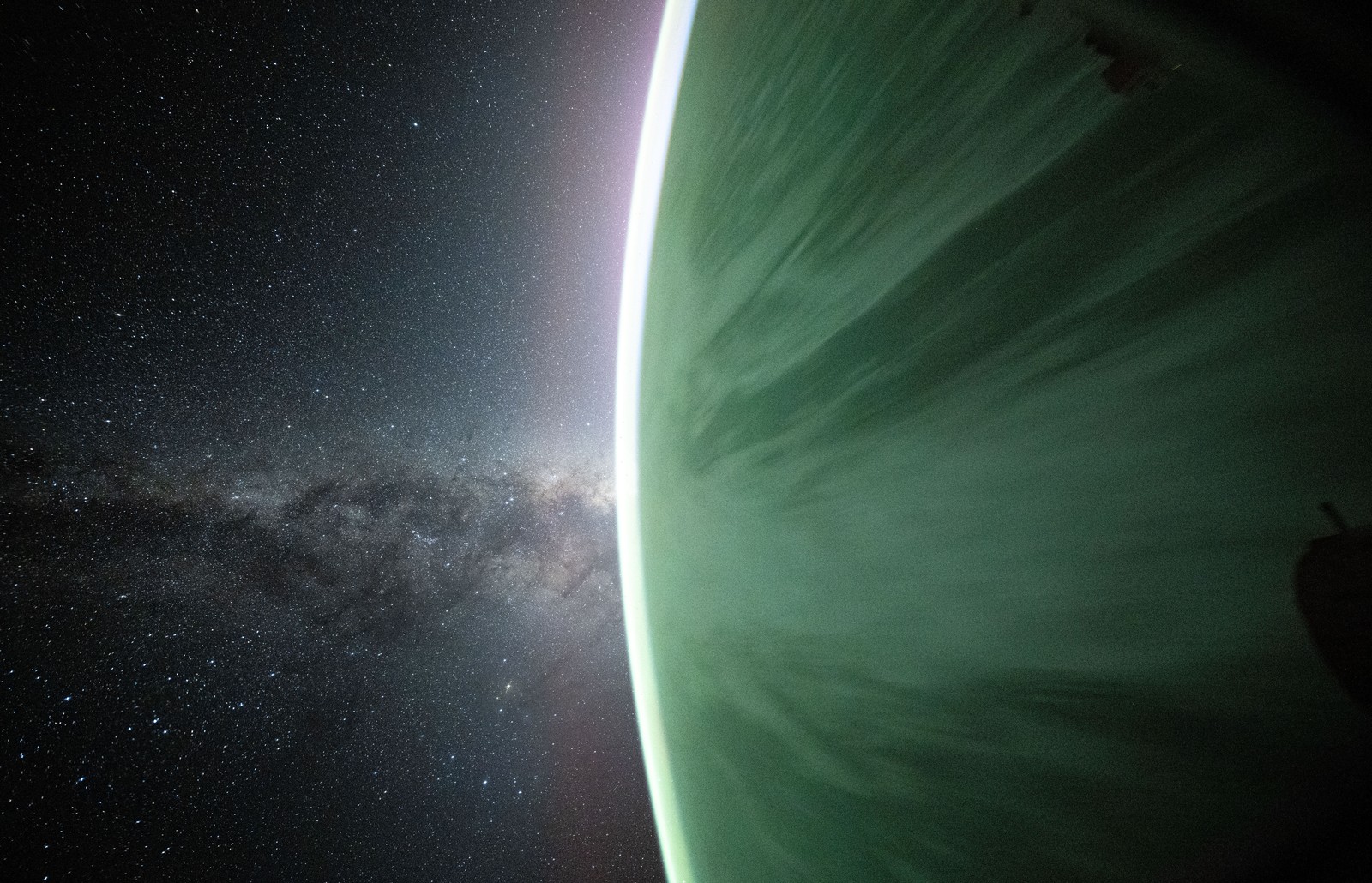 The Milky Way appears beyond Earth’s horizon, as the planet below appears beneath a slightly green glow.
