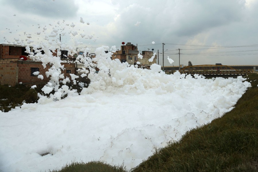 A small section of a river is completely covered by foam, some of it being blown by the wind into and over buildings on the shore.