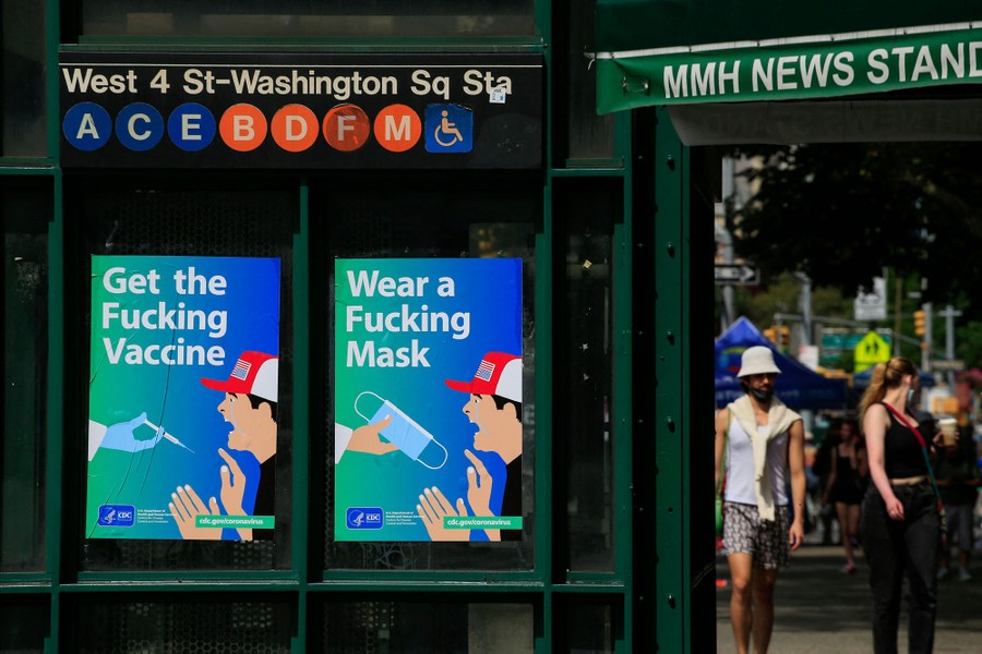 Two posters are seen outside a subway station, reading "Get the Fucking Vaccine" and "Wear a Fucking Mask."
