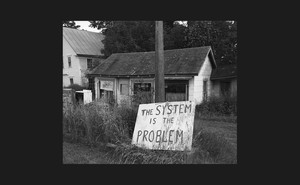 Black and white photo of ruined house with hand-painted sign "The System Is the Problem"