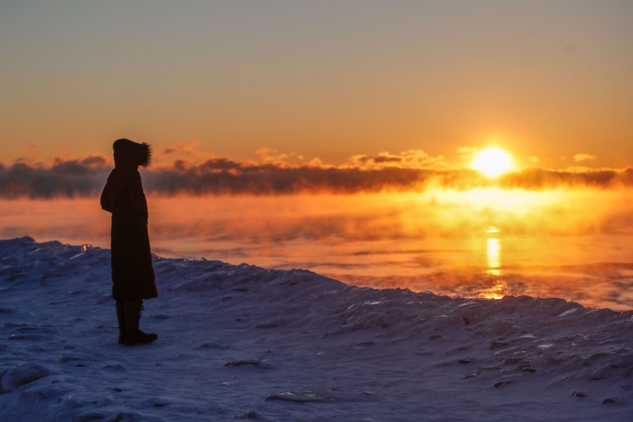 A person stands on a frozen lake shore at sunrise.