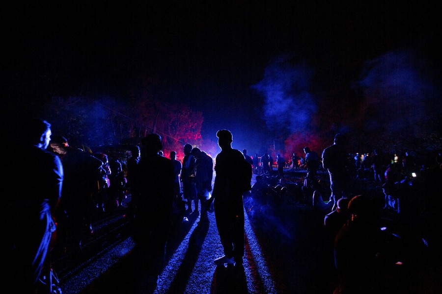 Dozens of people are seen in silhouette at night, standing outside in front of red and blue lights.