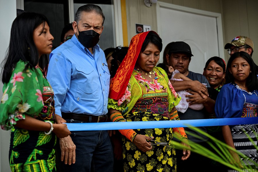 Several people stand in a small crowd, beside a house, as one person prepares to cut a blue ribbon with scissors.