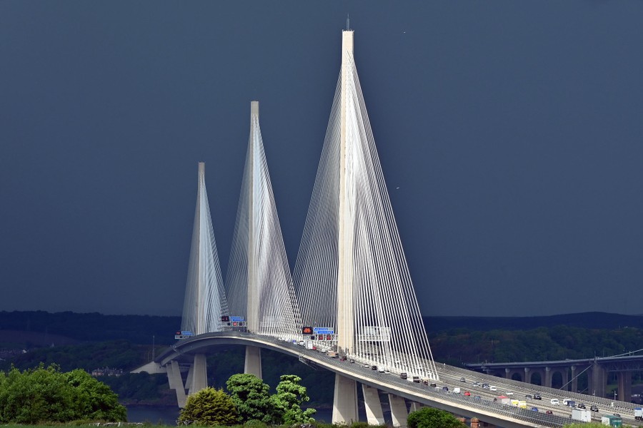 The tall supports and cables of a bridge shine in bright sunlight, backdropped by dark clouds.