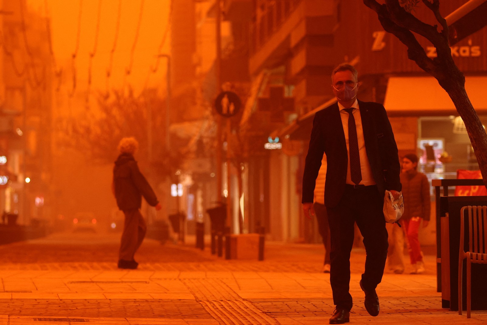 People walk through city streets, beneath a hazy orange sky, during a dust storm.