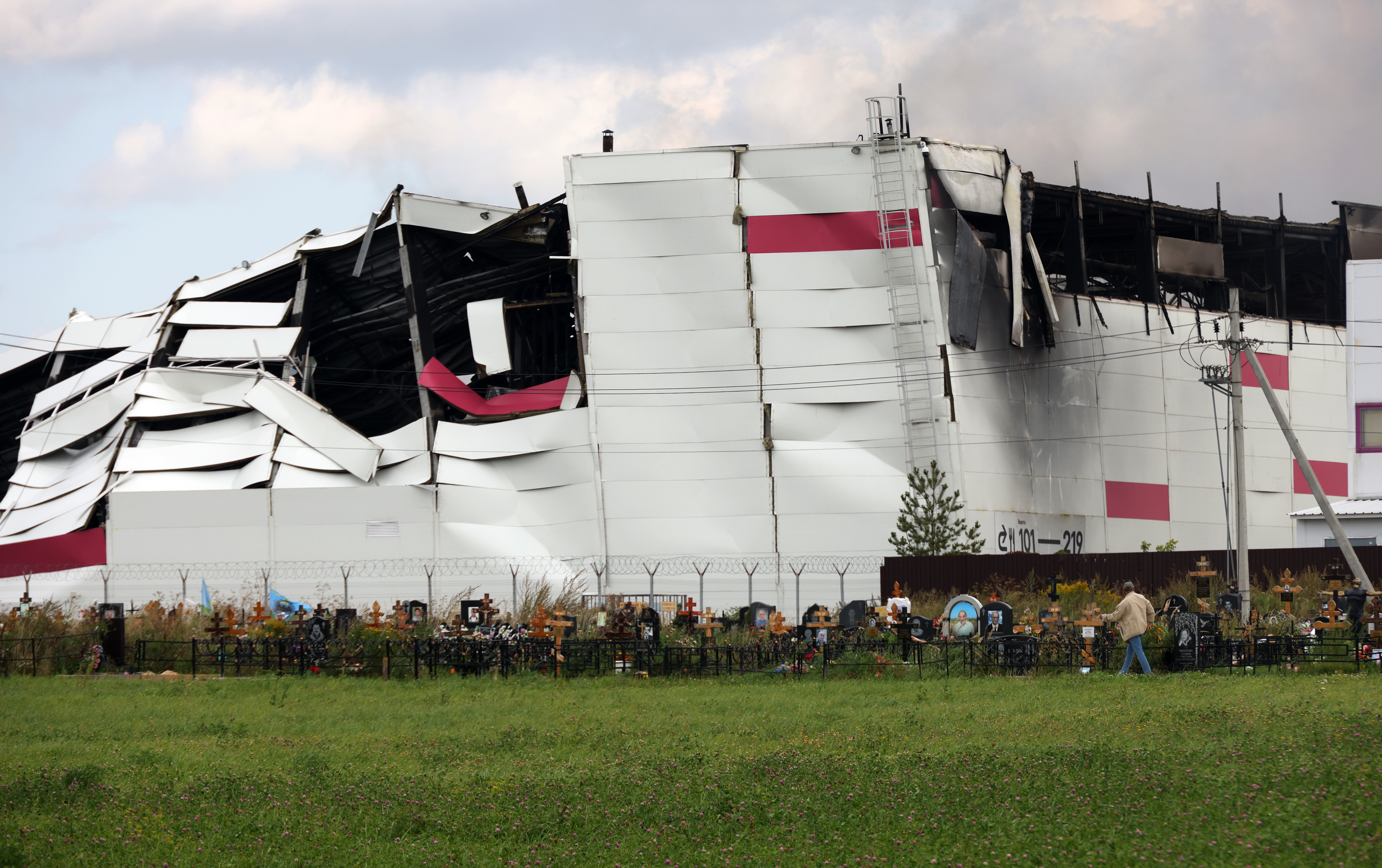 A person walks past gravestones that stand near the ruins of a burned-out warehouse in the background.