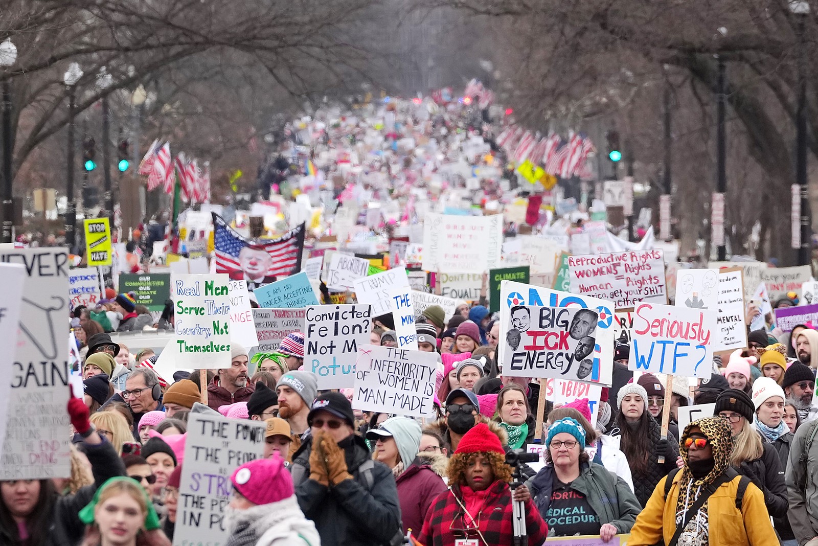 A large crowd of protesters carry signs with various messages, including 'The inferiority of women is man-made' and 'Real men serve their country, not themselves.'
