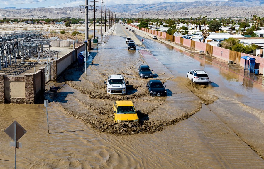 Several cars and trucks drive through knee-deep floodwater on a road.