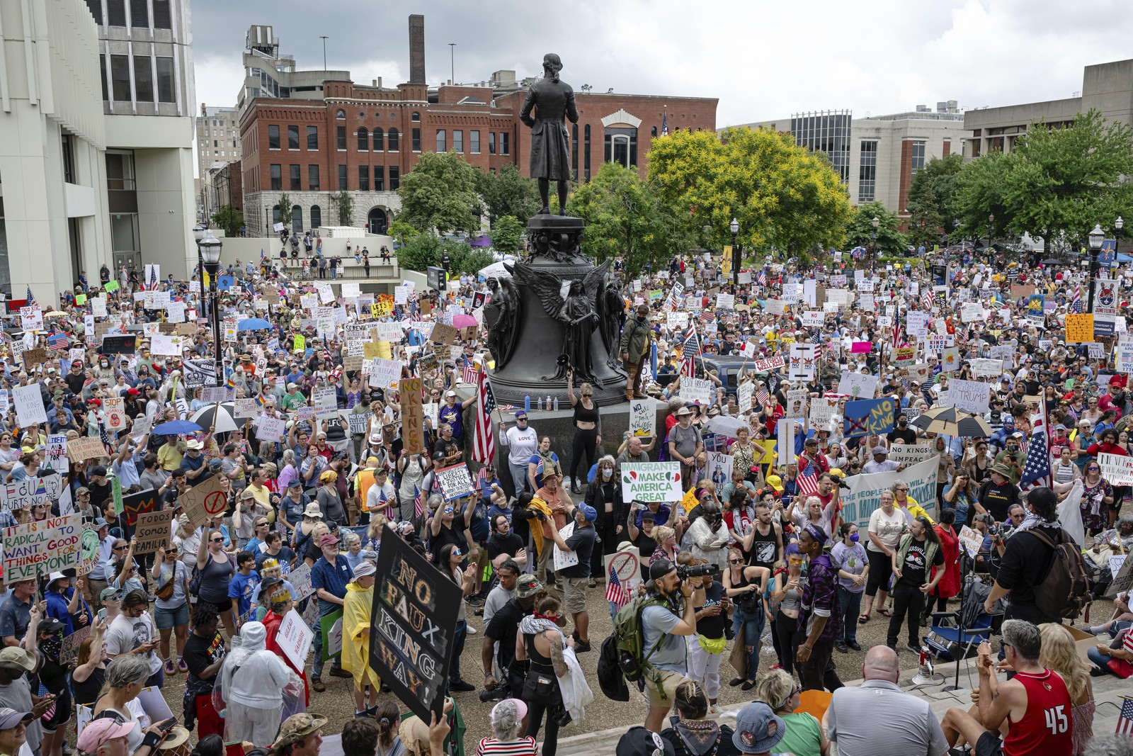 Protesters gather in a square, standing beside a statue.