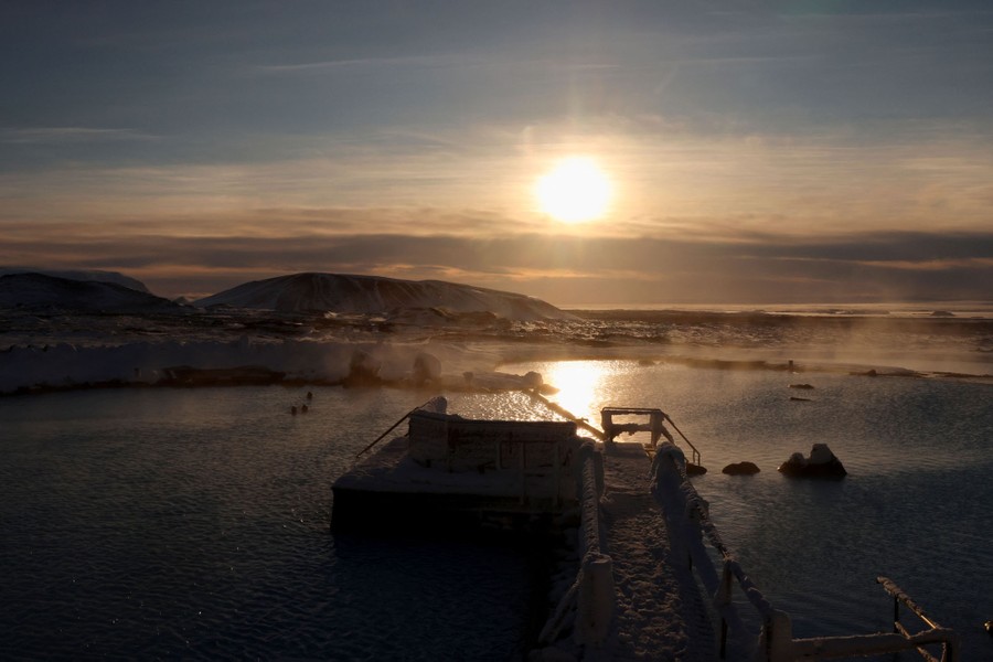 Several people swim in steamy pools outside, with snowy hills around.