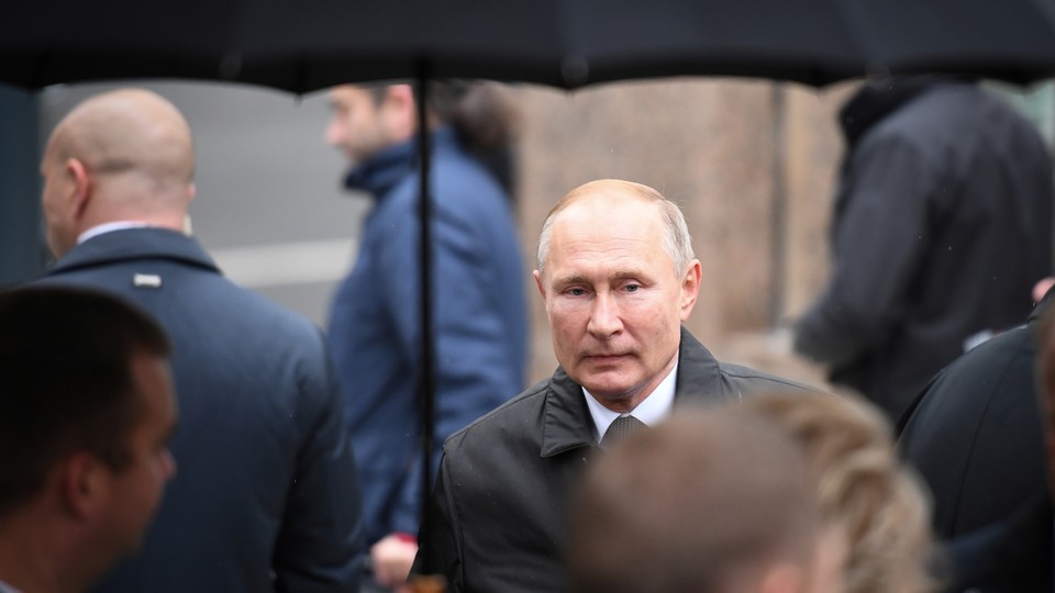 Vladimir Putin stands under an umbrella in the rain, surrounded by people facing the opposite direction.