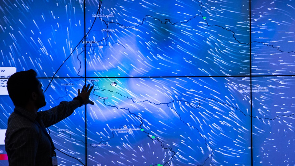 A meteorologist points to a big blue screen displaying wind data from hundreds of weather stations