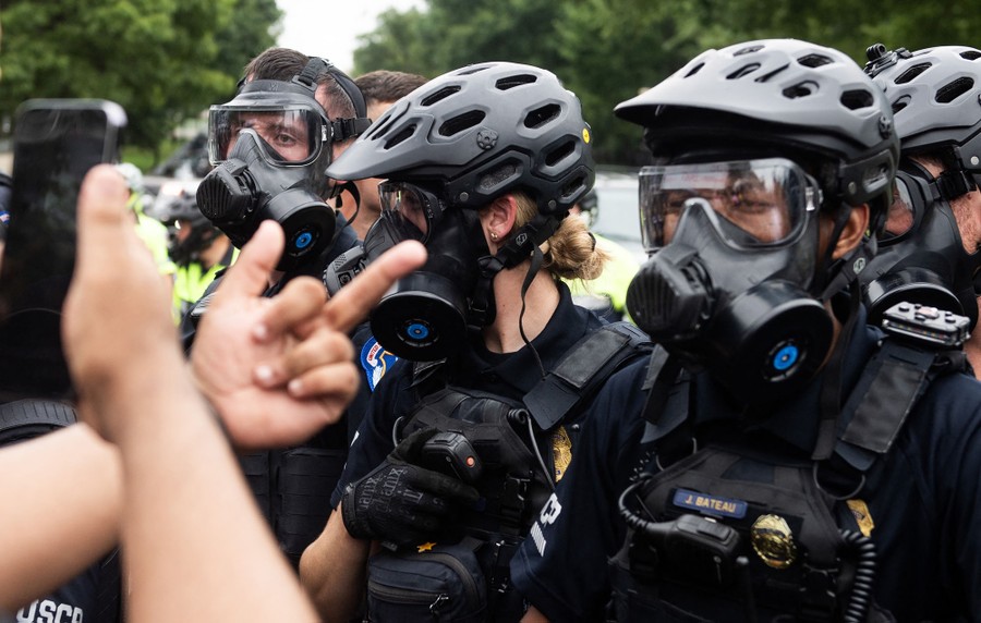 Riot police wearing helmets and gas masks stand in front of protesters. One of the protesters reaches out a hand, flipping off the police.