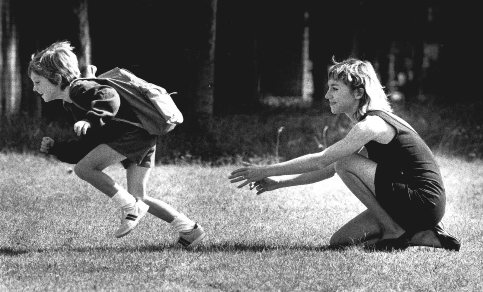 A boy carrying a backpack sprints gleefully from his mother's arms.