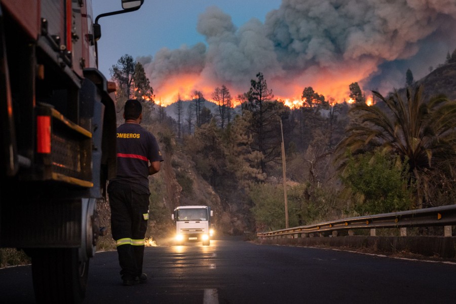 A firefighter stands on a road, looking up toward a hill where a wildfire burns.