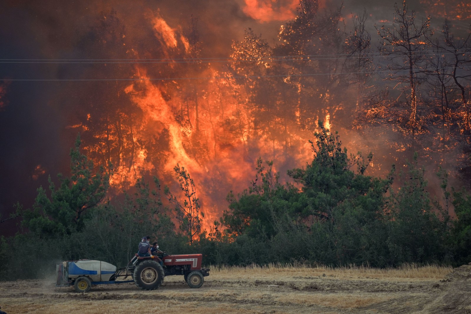 A farmer uses a tractor on a field, as a wildfire burns the forest in the background.