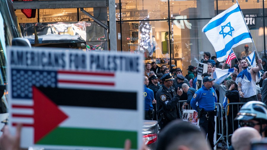 Protesters stand on a street, separated by police and barriers, flying flags in support of either Israel or Palestine.