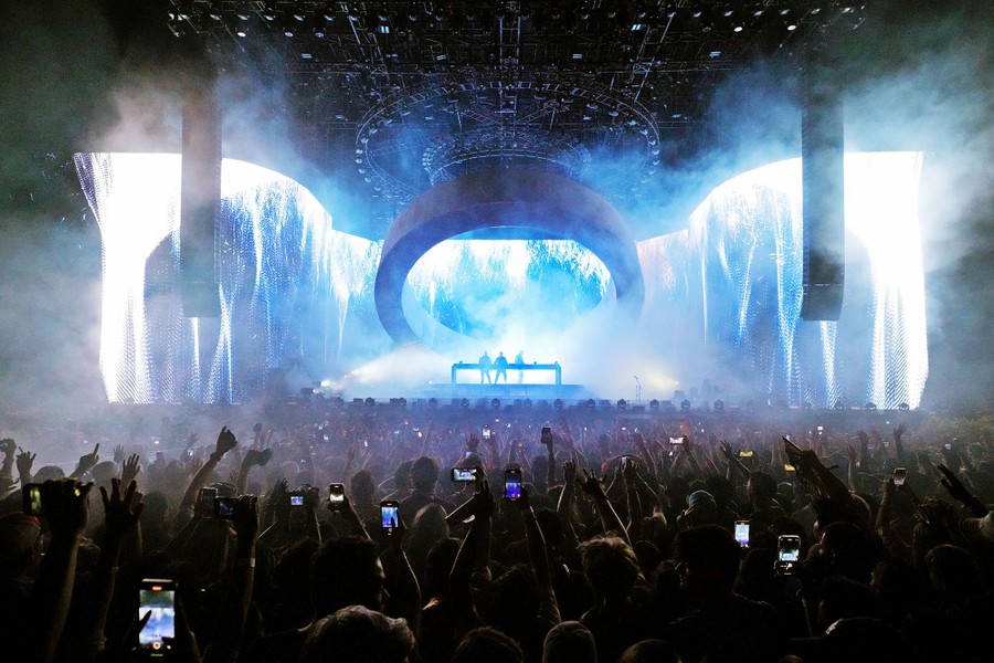 A large crowd enjoys a musical performance, with the stage brightly lit by a video display and stage lighting.