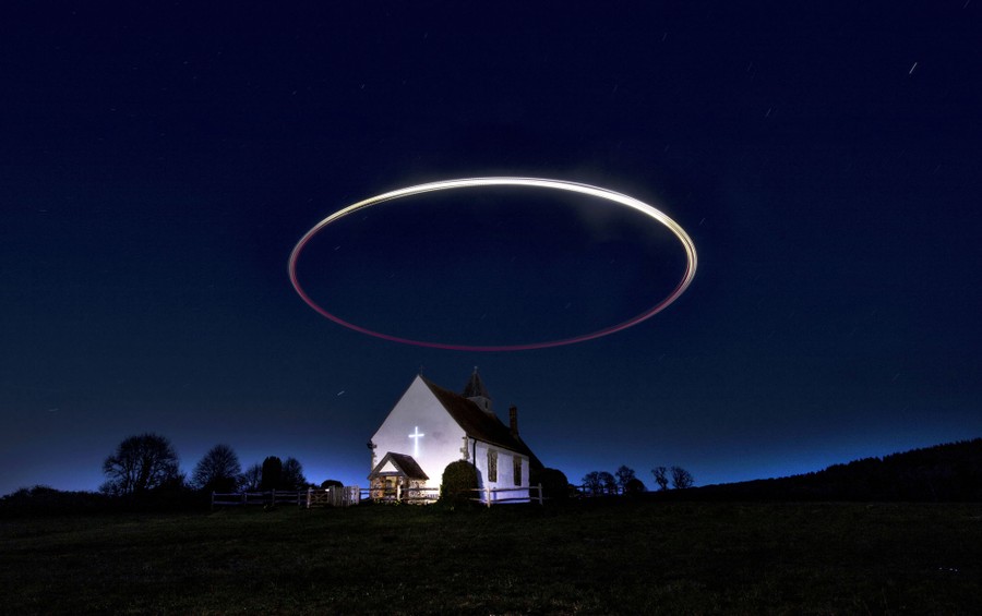 A circle of light is seen in the sky above a small church in a field.
