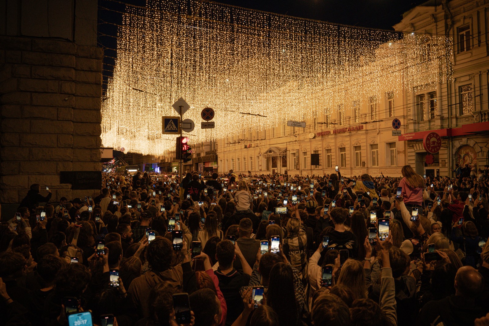 A crowd of people hold their phones up to take pictures of a city plaza lit by many strings of hanging lights.
