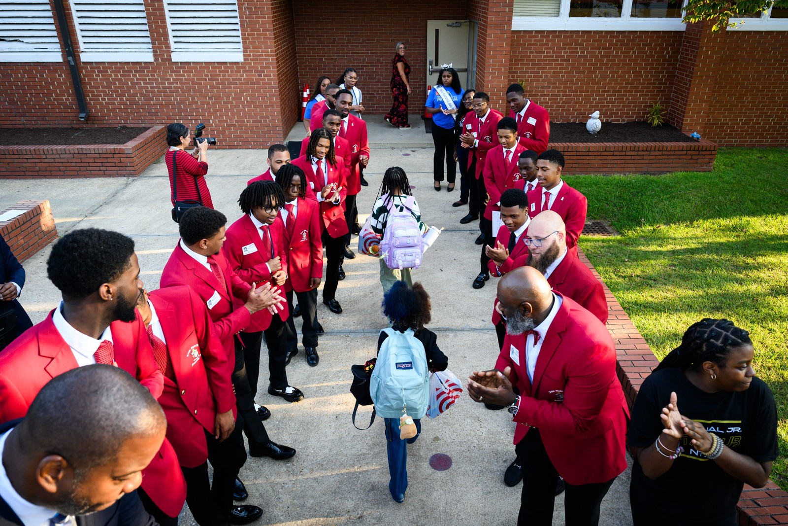 Two lines of people stand on either side of a path, clapping and welcoming young students into a school.