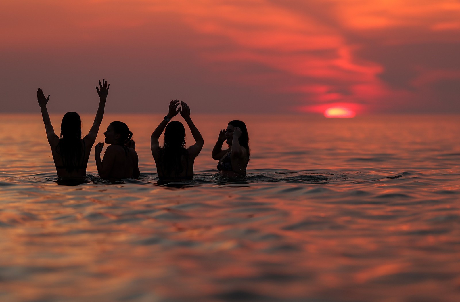 Four women stand waist-deep in calm water at sunset.