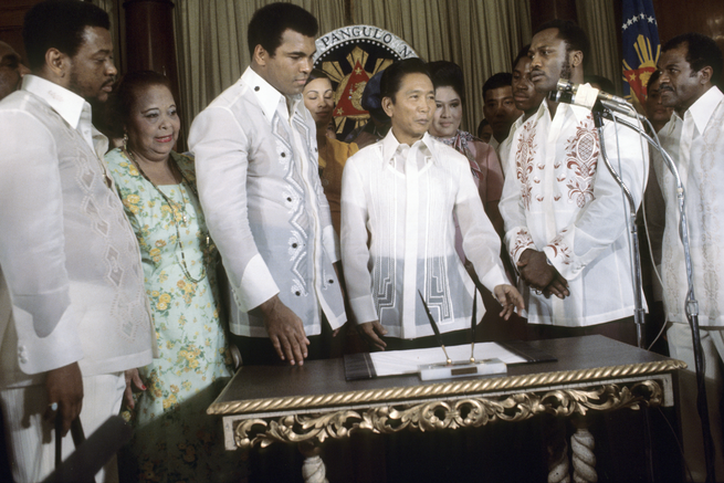 color photo of 4 men and 1 woman, all the men in different patterned white embroidered long-sleeve dress shirts, standing in a row behind an ornate desk in front of state seal, with microphone