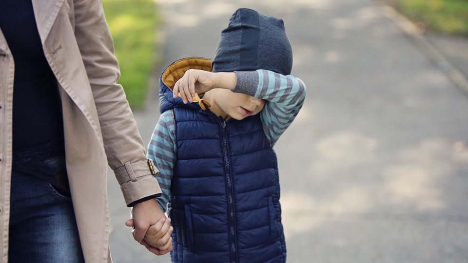 A boy holds his arm in front of his face while holding hands with a woman