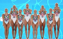 Eight swimmers wearing swimsuits with cat faces printed on them perform in a line beside a pool.