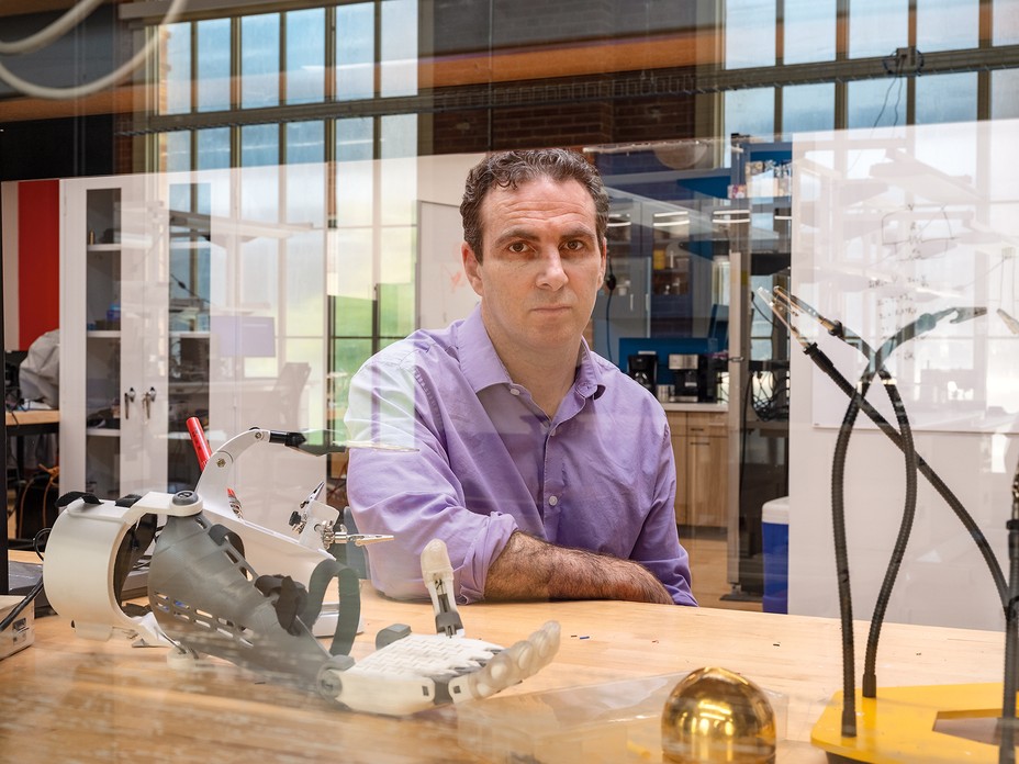 James sitting at wood-topped table behind reflective glass, with clamps and robotic hand on table in front of him 