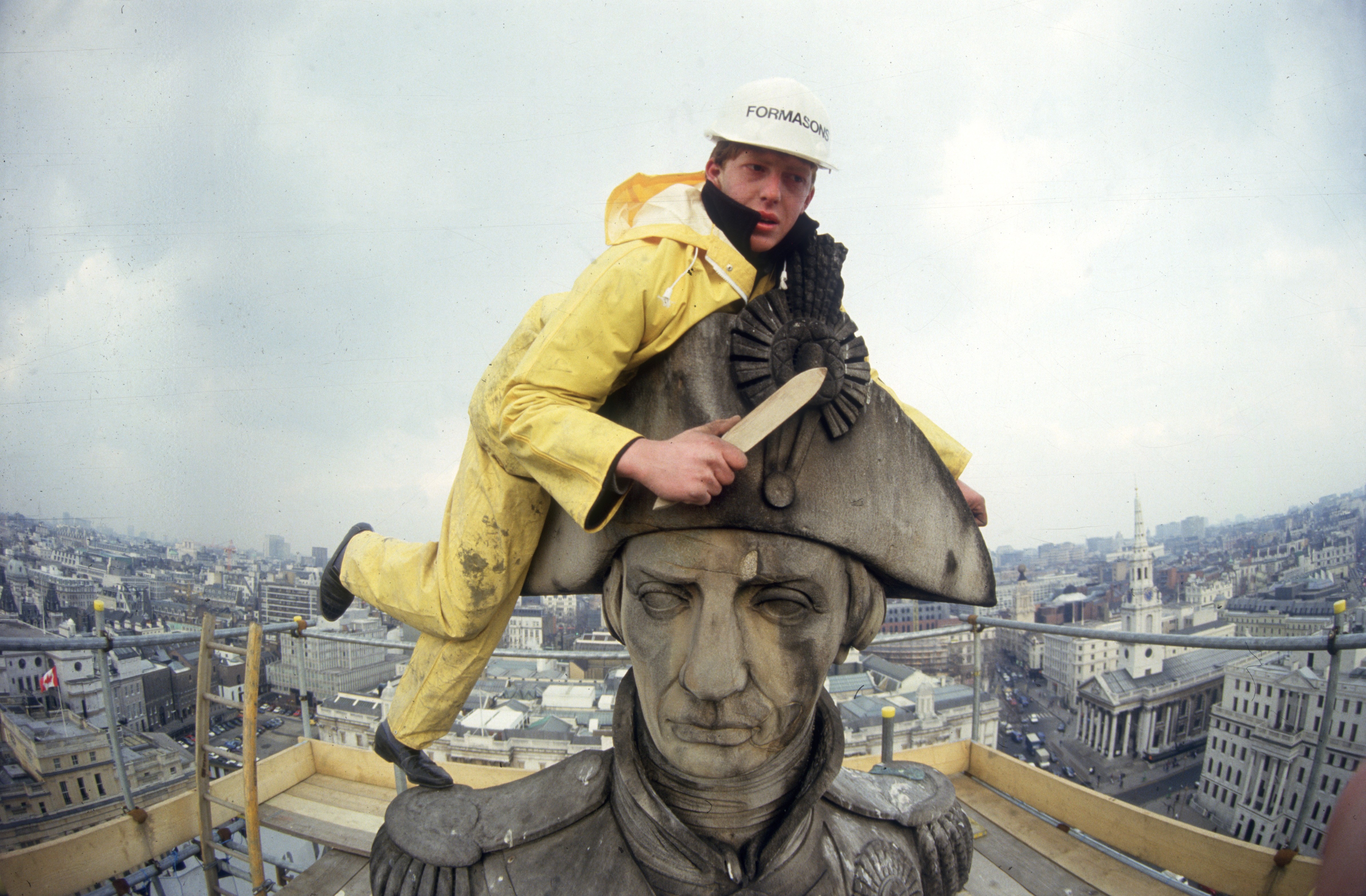 A worker with a brush leans on the head of a statue, high above London, on scaffolding.