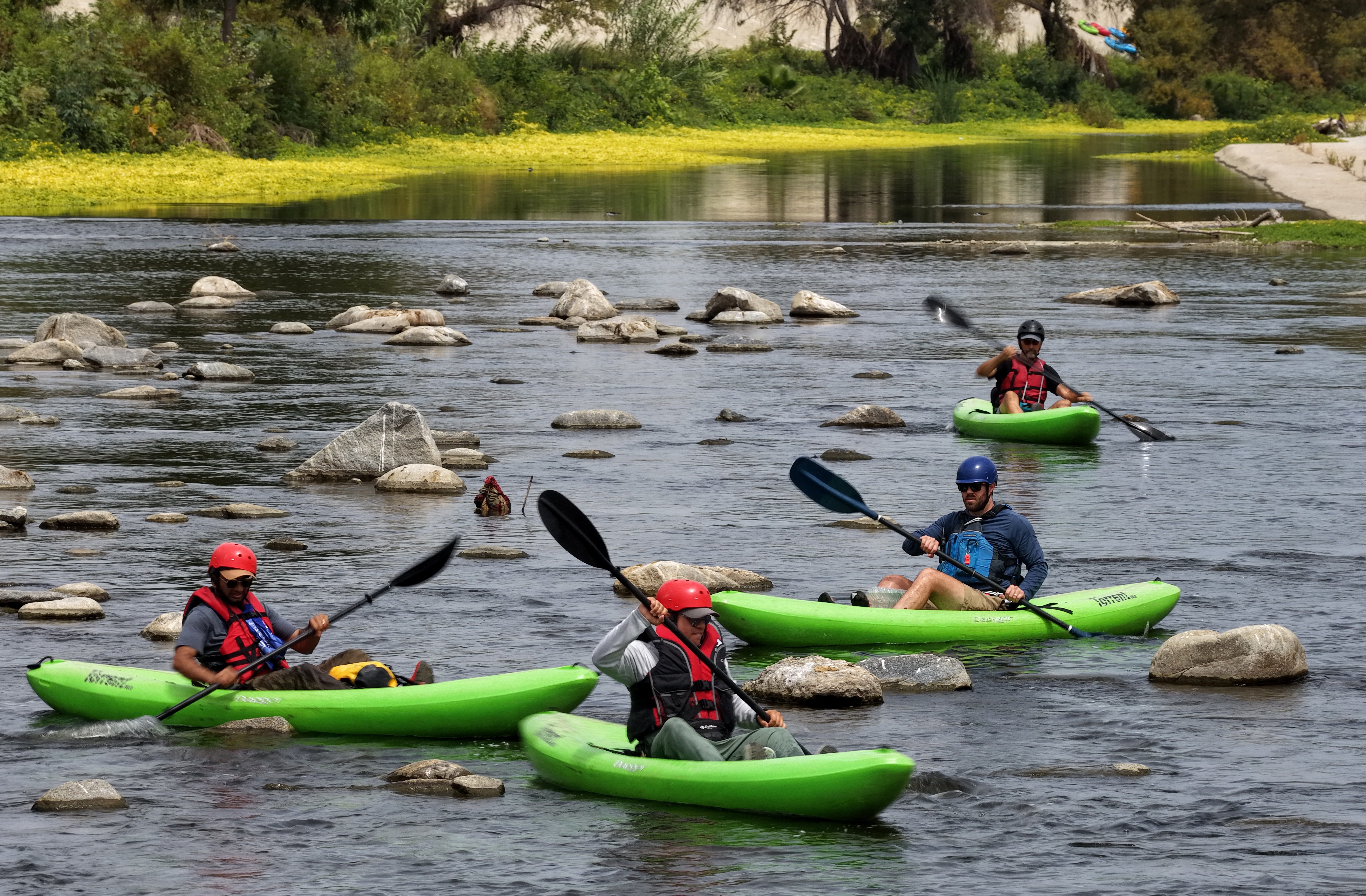 Kayakers navigate past small rocks in a shallow river.