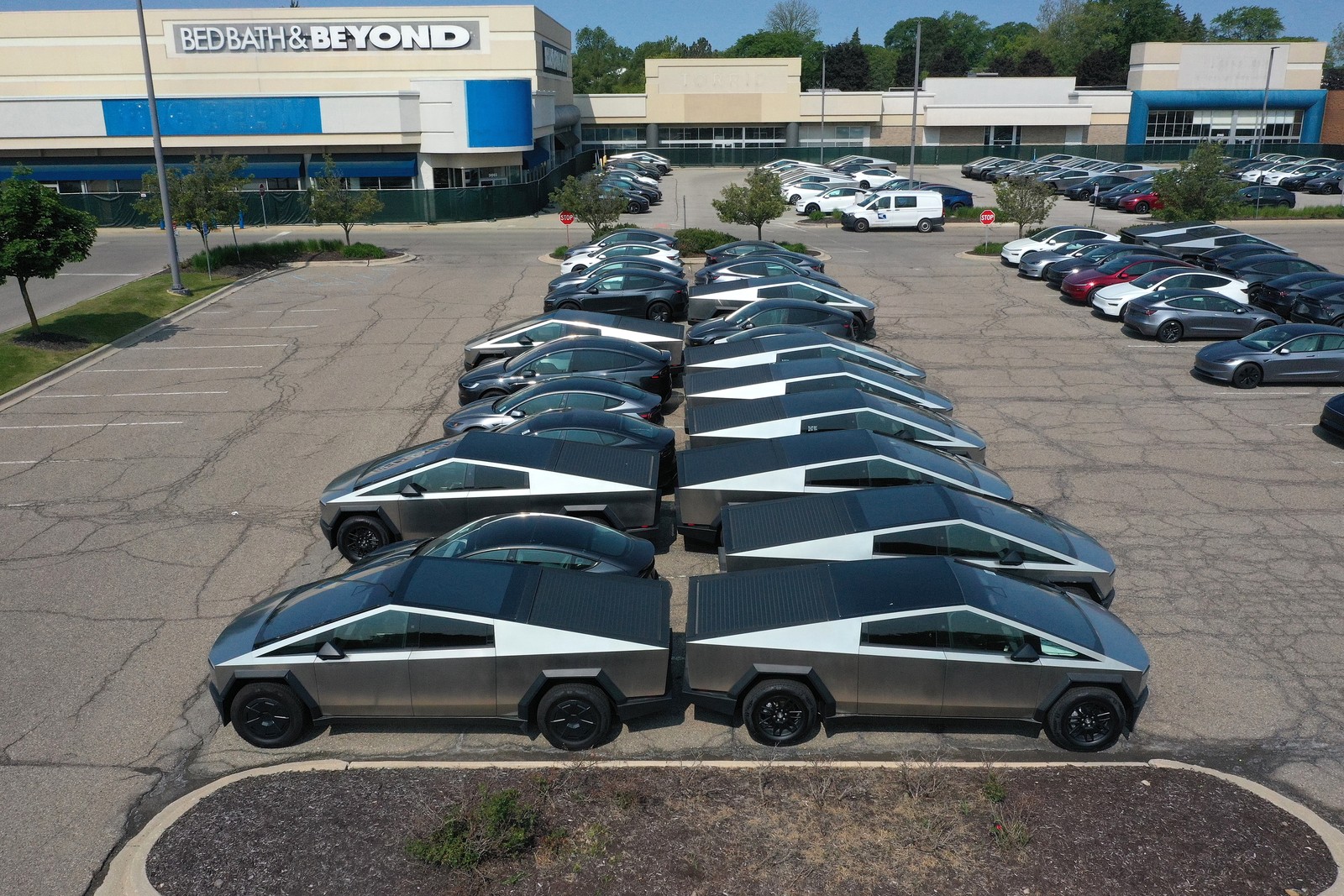 An elevated view of dozens of cars, including many cybertrucks, sitting in the parking lot of a closed mall.