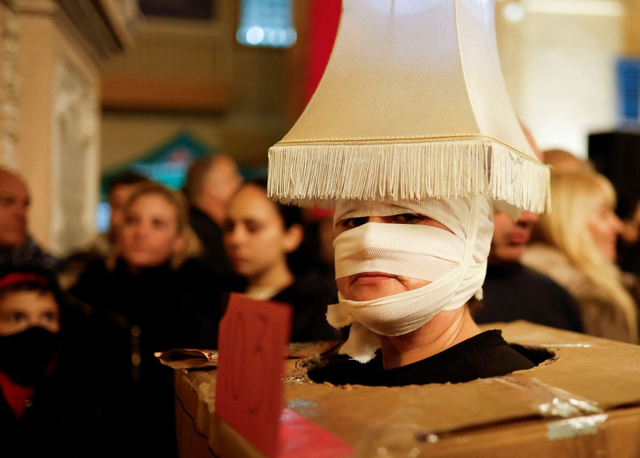 A costumed Carnival participant wears a large cardboard box over their torso, a bandage around their head, and a lampshade as a hat.
