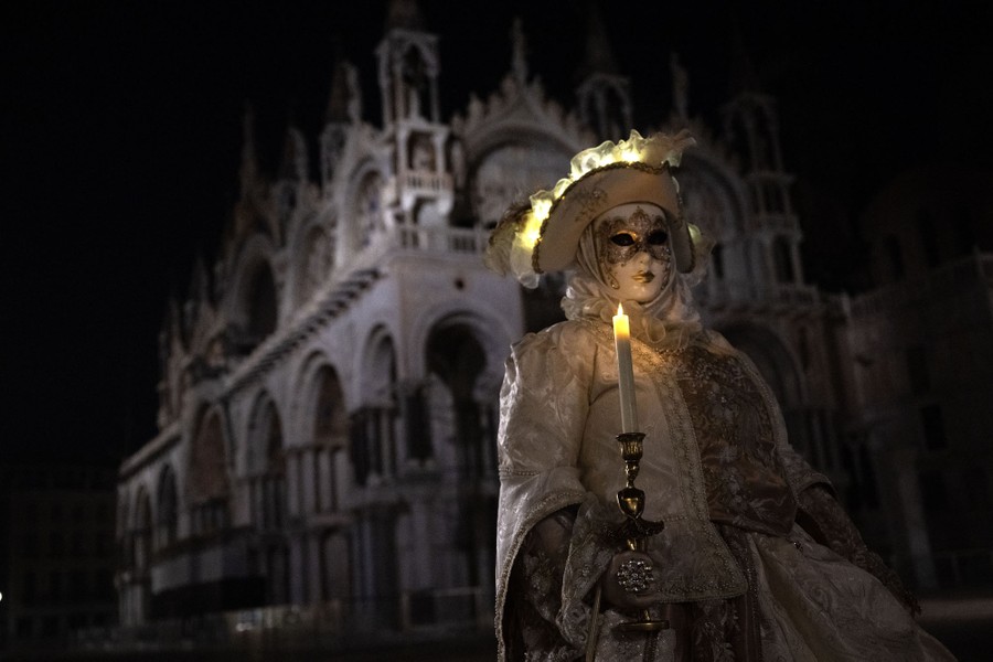 A person wears a mask and period costume, holding a candle, in front of an ornate building in Venice.