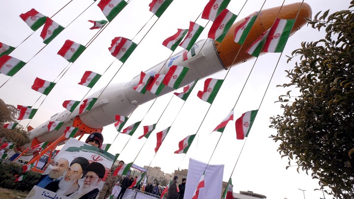 A boy holding a placard with pictures of President Hassan Rouhani, Ayatollah Ruhollah Khomeini, and Ayatollah Ali Khamenei, poses for camera in front of a model of Simorgh satellite-carrier rocket with Iranian flags everywhere.