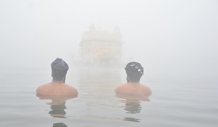 Two people sit in water up to their shoulders, looking toward a temple structure nearby, engulfed in smog.