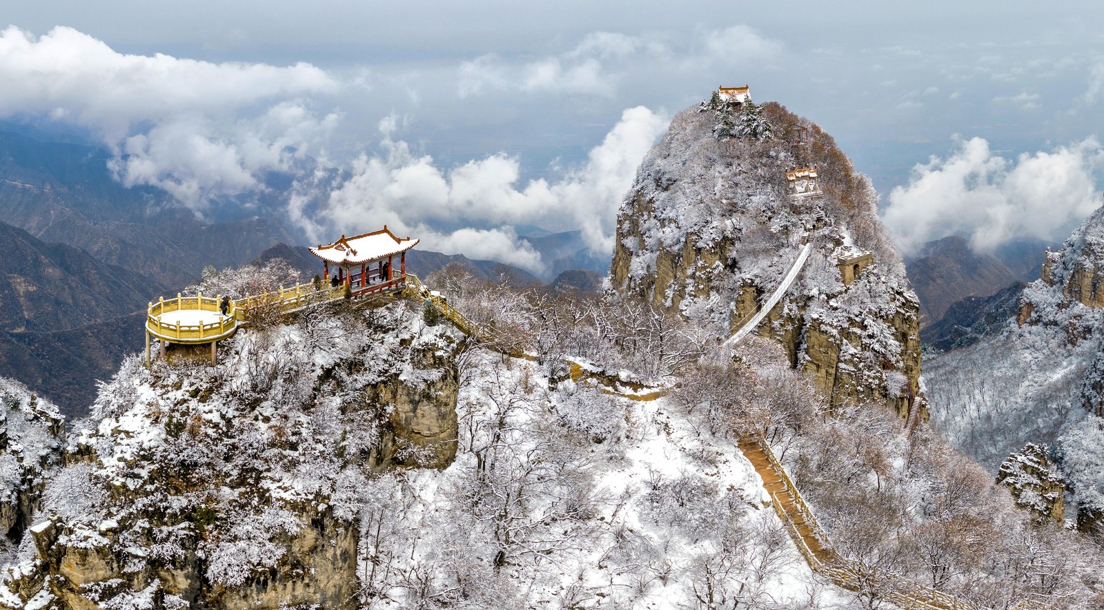 An aerial view of a steep set of snow-covered mountaintop rock formations, dotted with small temples and pathways.