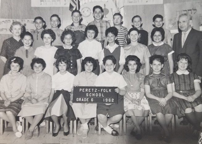 A black and white photo of a class of kids with a teacher on the far right. Two kids in the front row hold a sign that reads "Peretz-Folk School Grade 6 1962"
