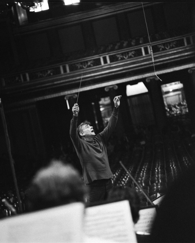 black-and-white photo of Leonard Bernstein onstage, arms raised overhead with baton, with theater balcony in background and sheet music and musicians' heads in foreground