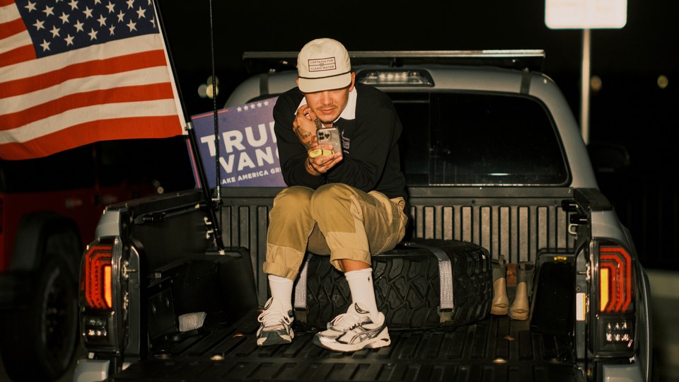 Young man sitting in the back of a pickup truck, checking his phone, with a Trump campaign sign and the American flag behind him