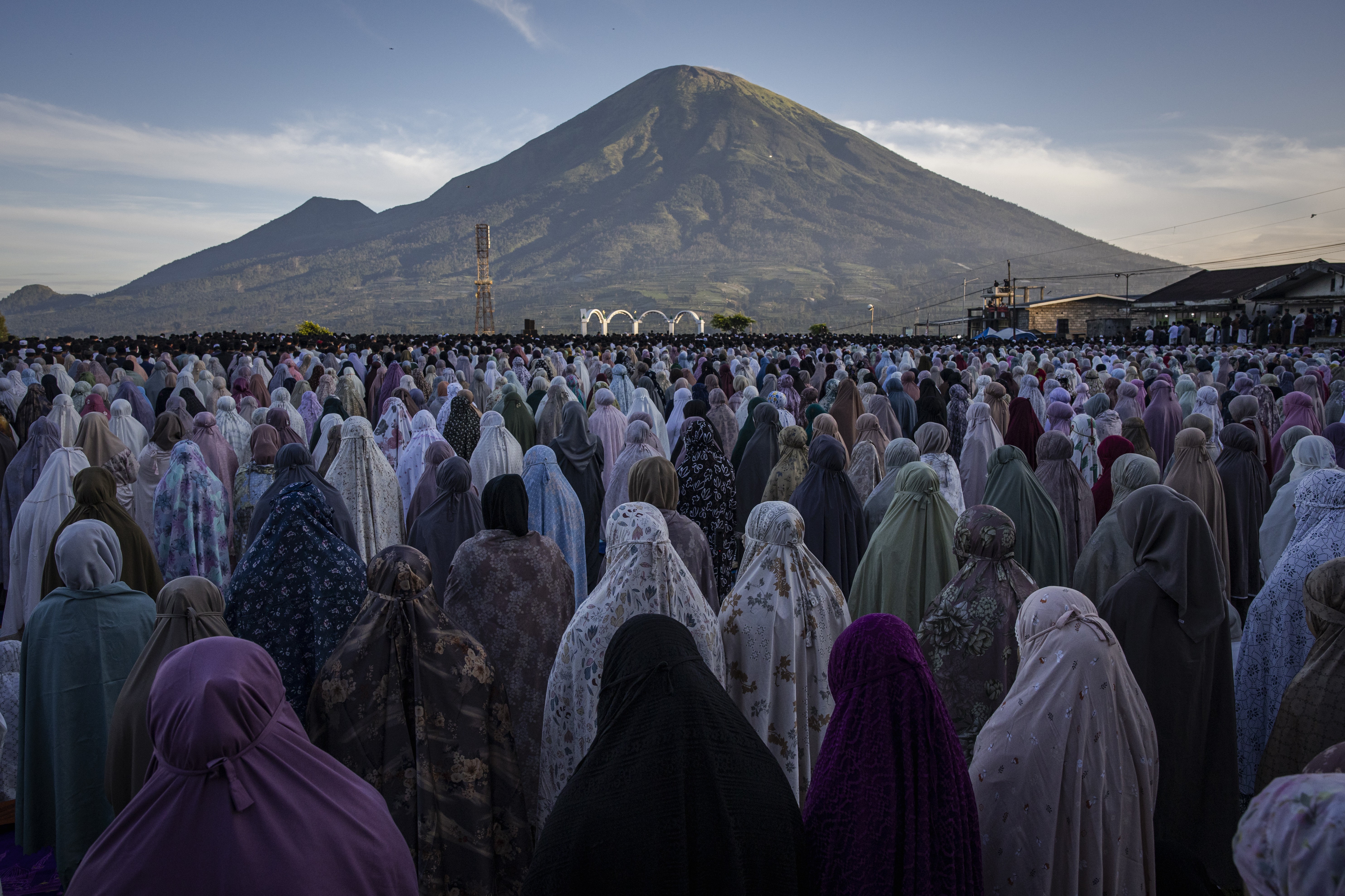 A large group of veiled Muslim women stand together beneath a mountain during a prayers.