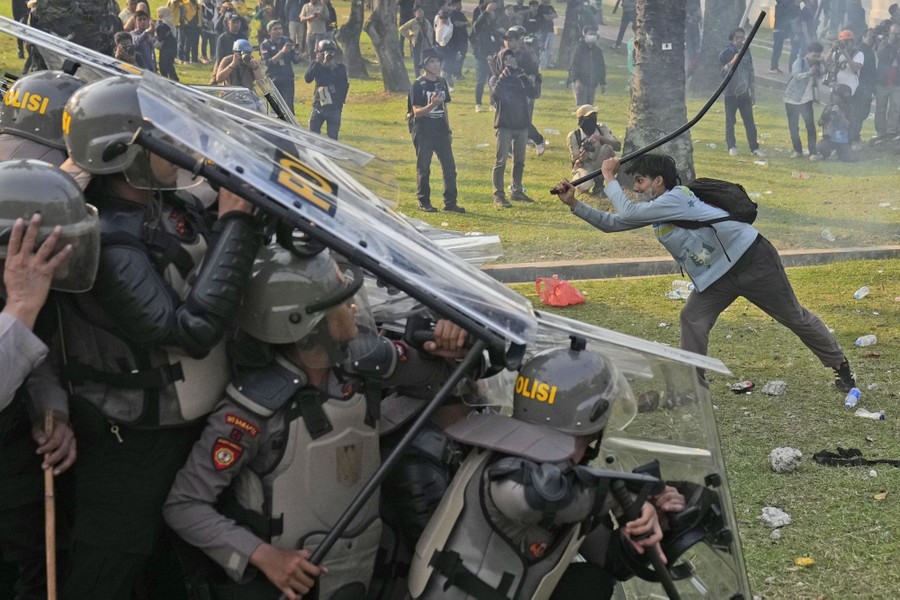 Watched by a crowd of onlookers, a protester uses a pole to hit the shields of many riot police who have clustered together in a defensive stance.