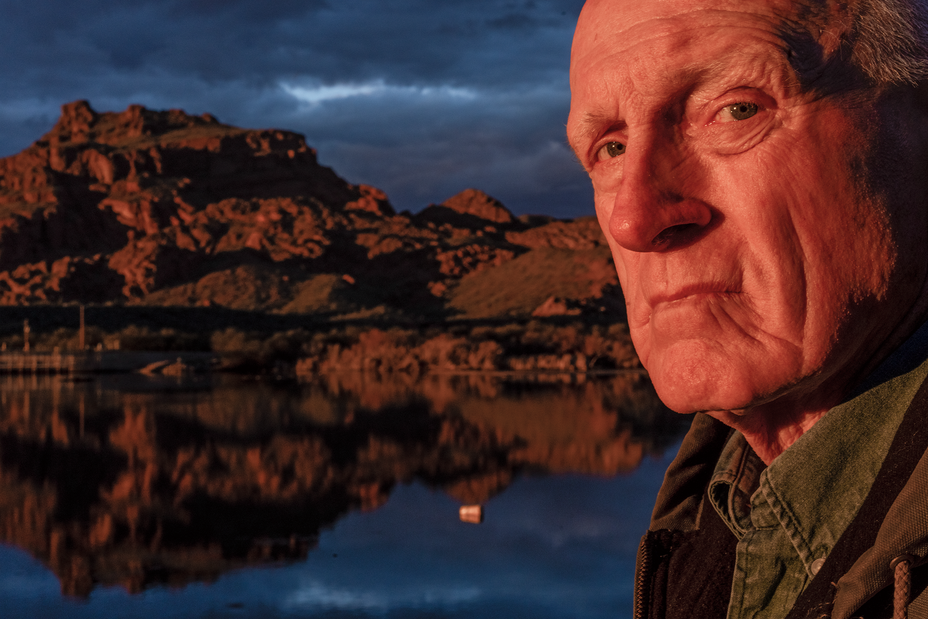 photo of man's face in reddish sunlight with water, rocky landscape, and dark clouds behind
