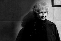 A black-and-white portrait of Alice Munro, standing against a wall and gazing to the right