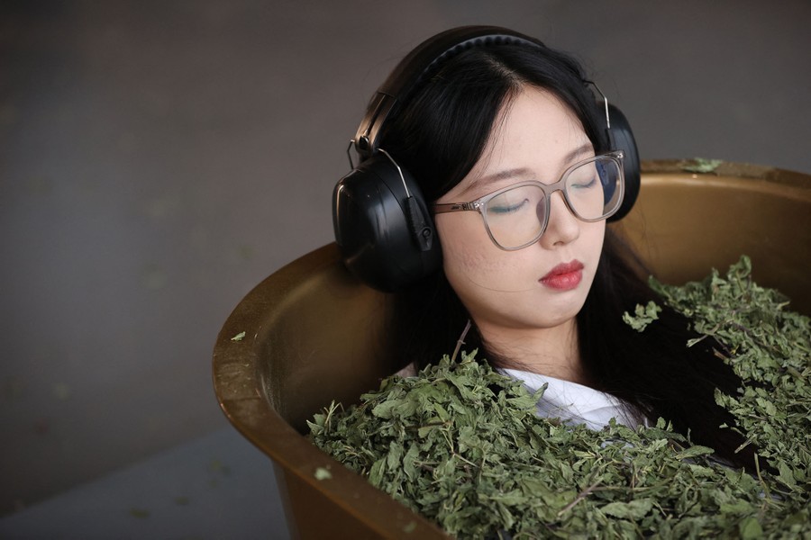 A person lies in a tub filled with dried leaves, wearing headphones.