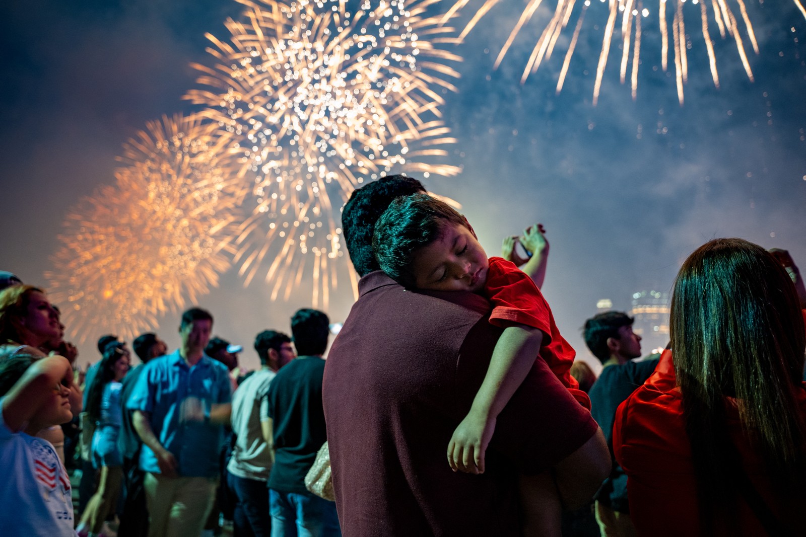 A man holds a sleepy young boy in his arms as fireworks erupt overhead.