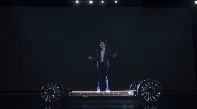 The writer Malcolm Gladwell stands on an electric-car chassis and shrugs 