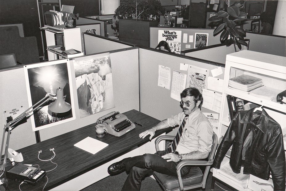 black and white photo of office cubicles with man in rolled shirtsleeves, tie, and dark sunglasses sitting at desk in foreground with typewriter